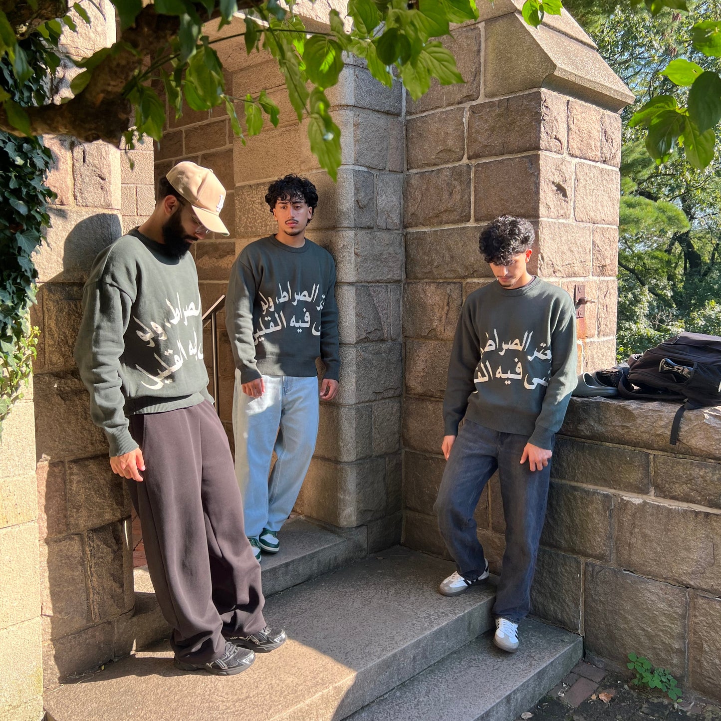 Three people standing on stone steps with a stone wall and greenery in the background. Green sweater. Knitted