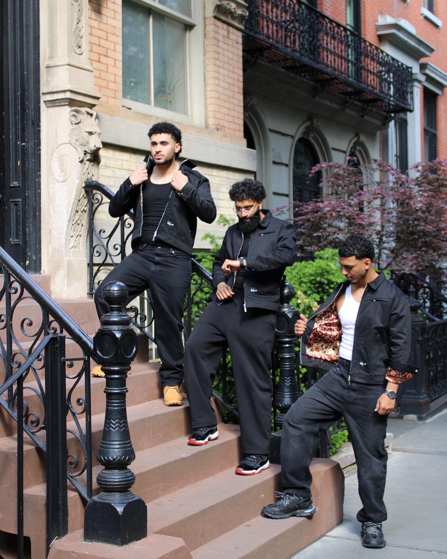 Three men in black outfits standing on a city street with brick buildings and iron railings.