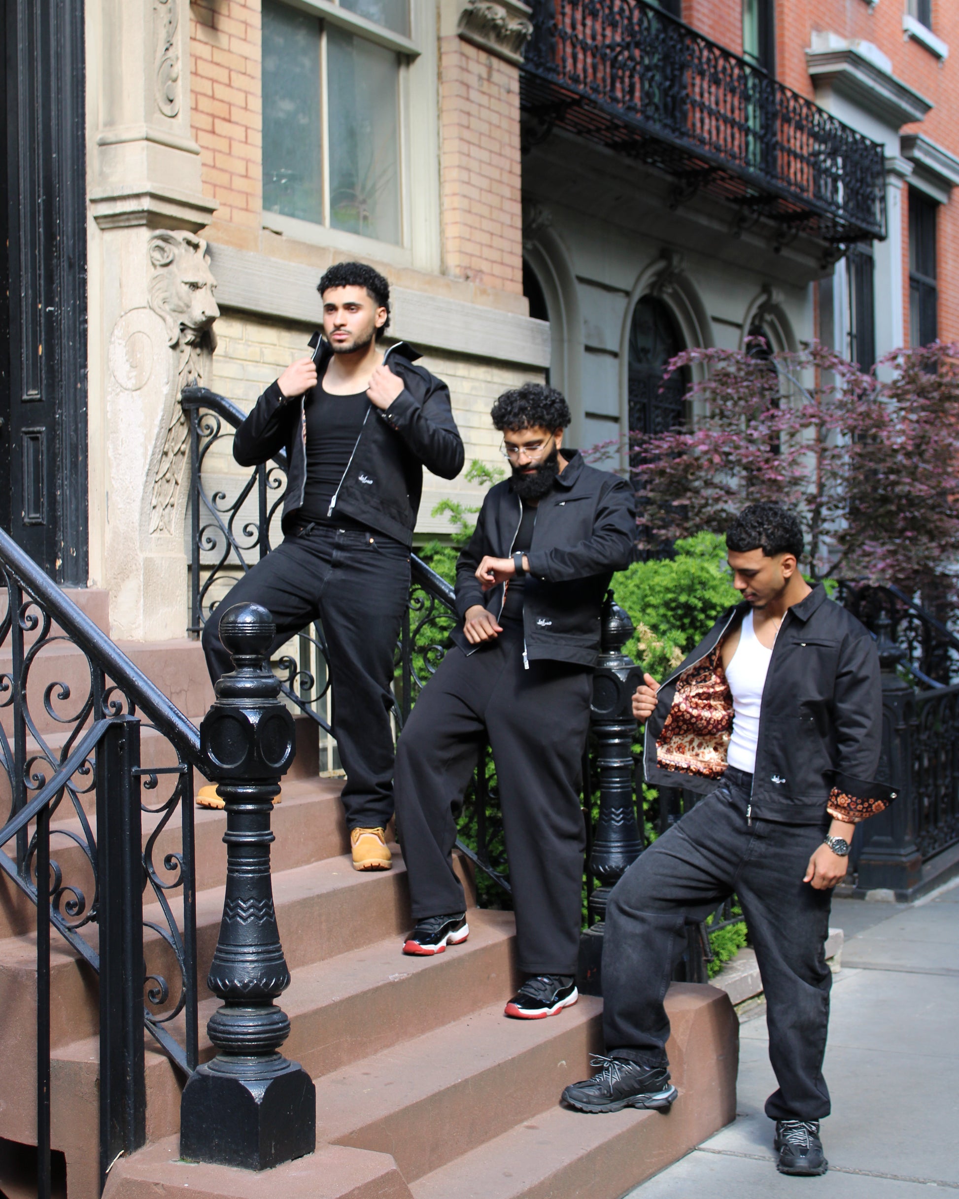 Three men in black outfits standing on a city street with brick buildings and iron railings.