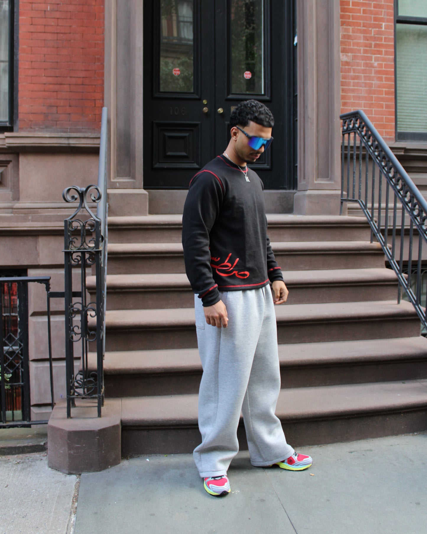 Man standing on a set of stairs in front of a brick building wearing a black sweater with red designs, light gray pants, and colorful sneakers.